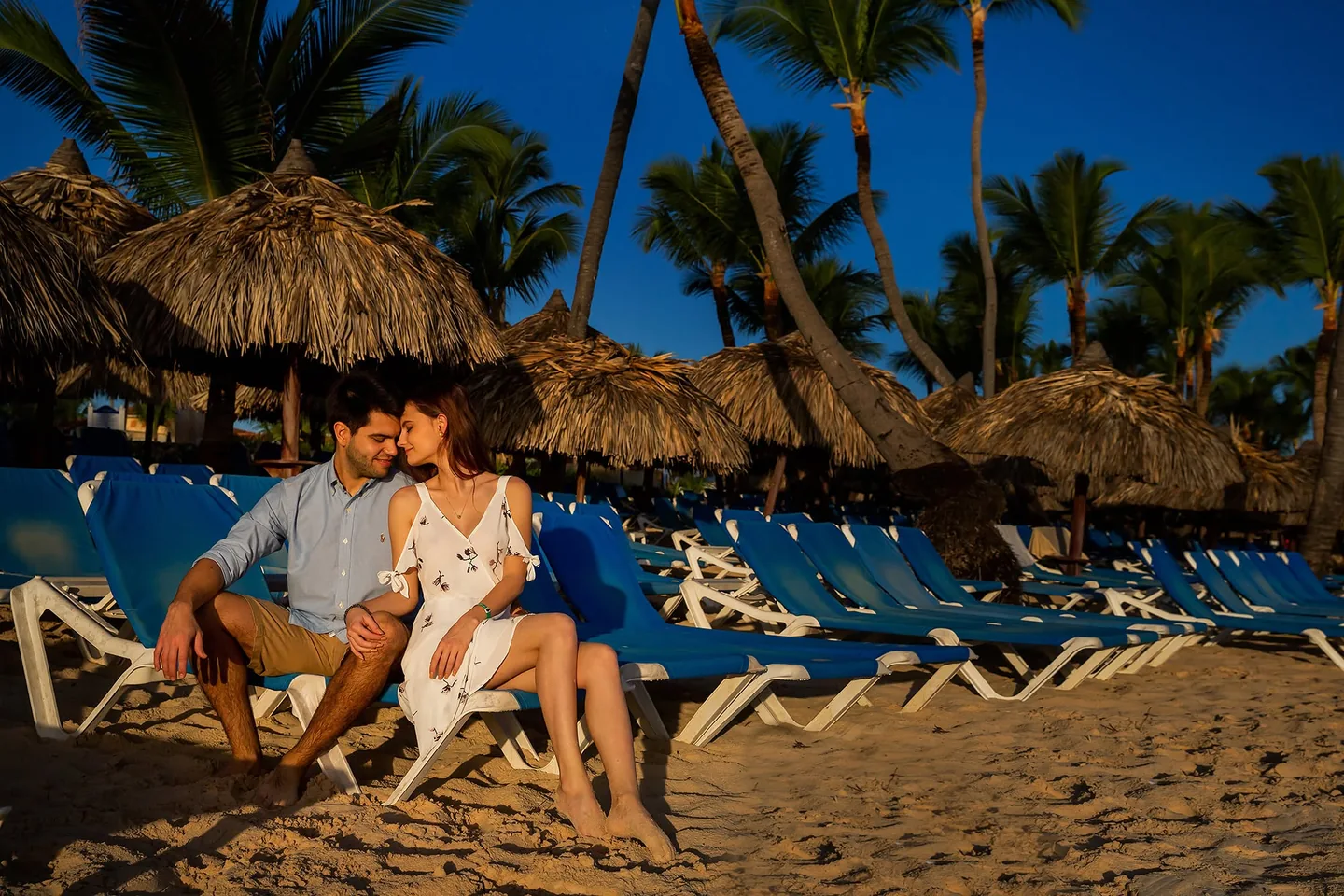Couple sitting close together on beach loungers beneath palm trees