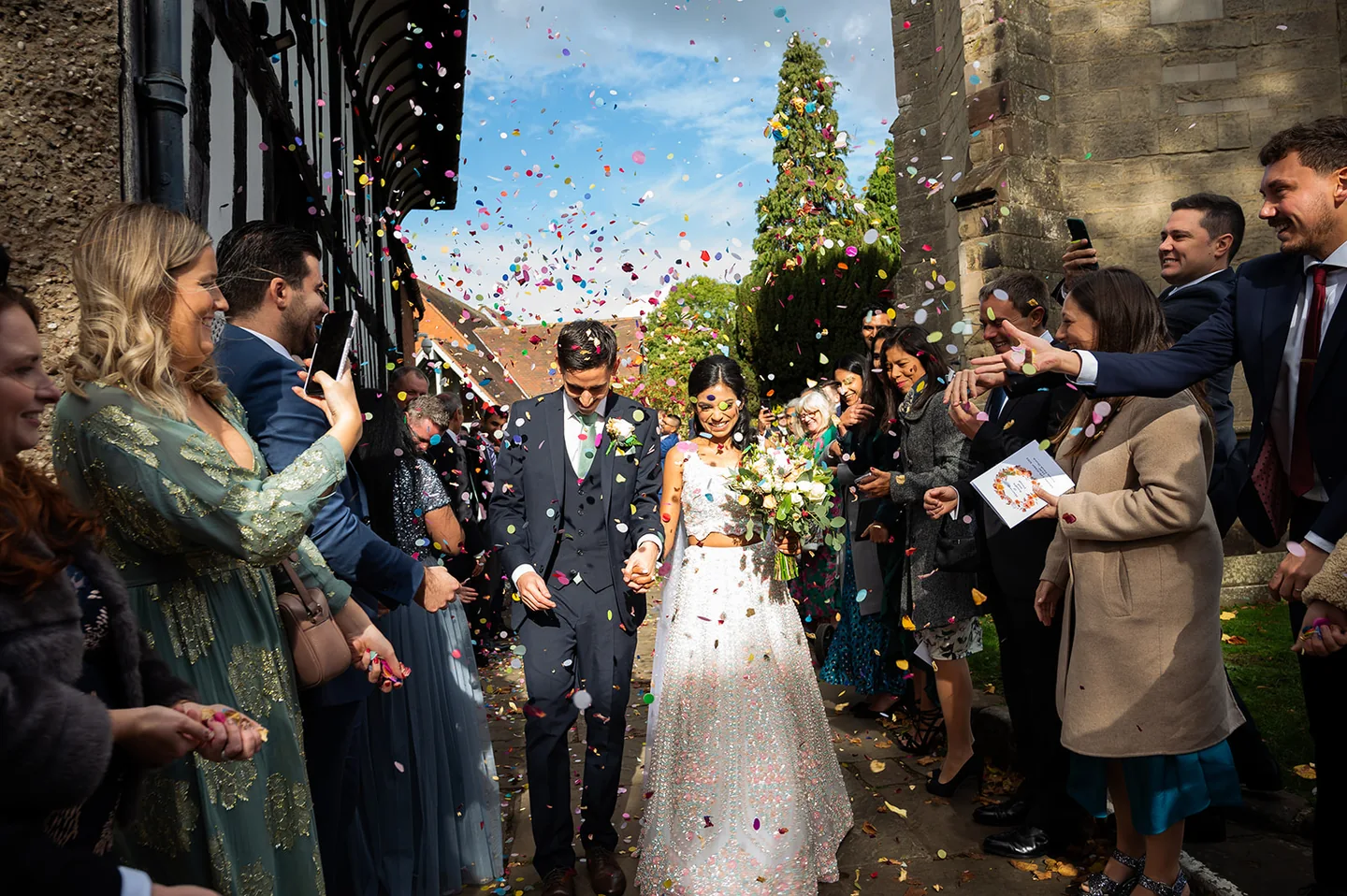 Newlyweds holding hands beneath colourful confetti outside a stone church