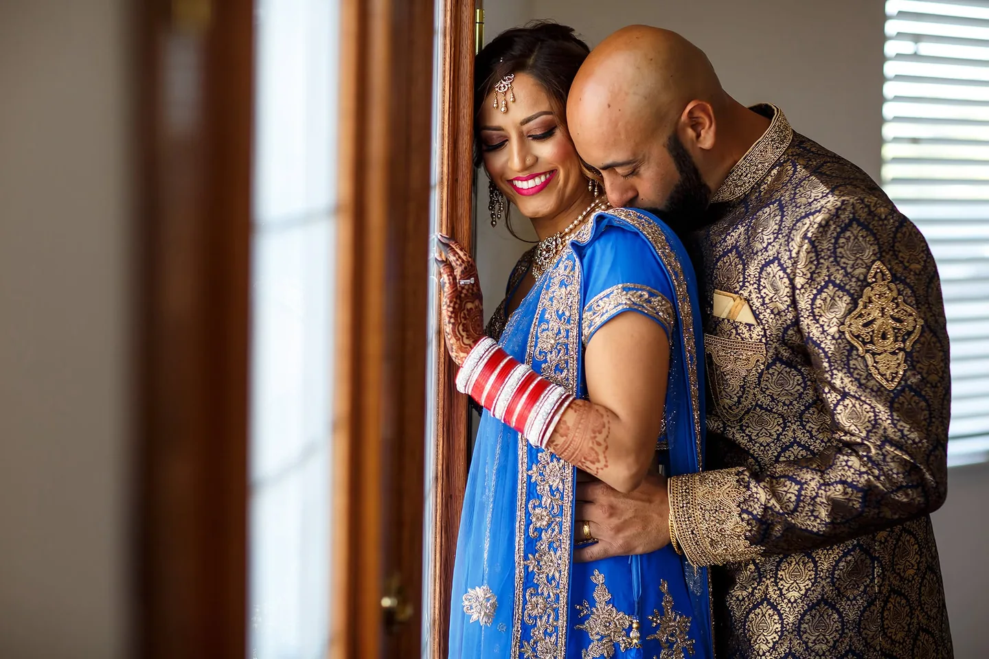 Wedding couple in blue and gold embracing beside a sunlit window