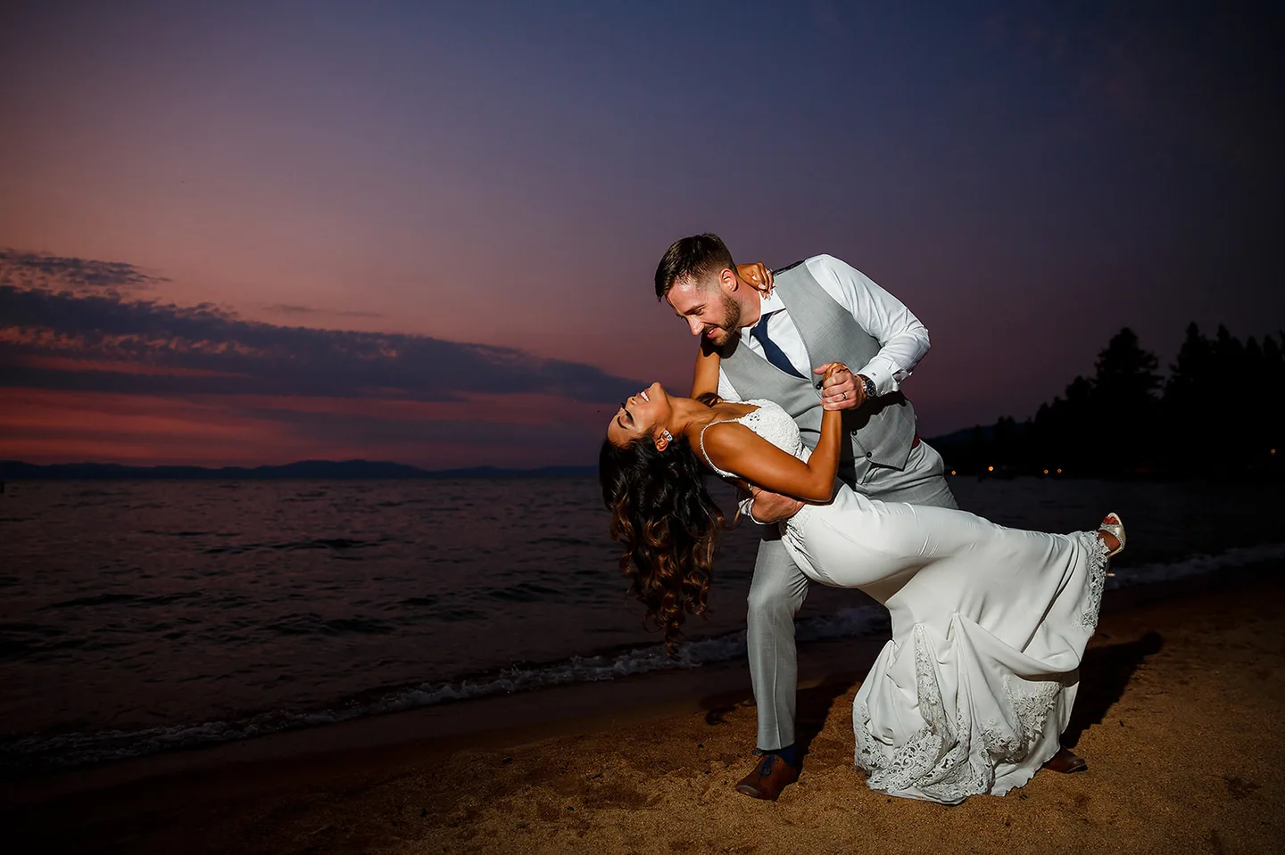 Groom dipping his bride beside the water beneath a purple evening sky