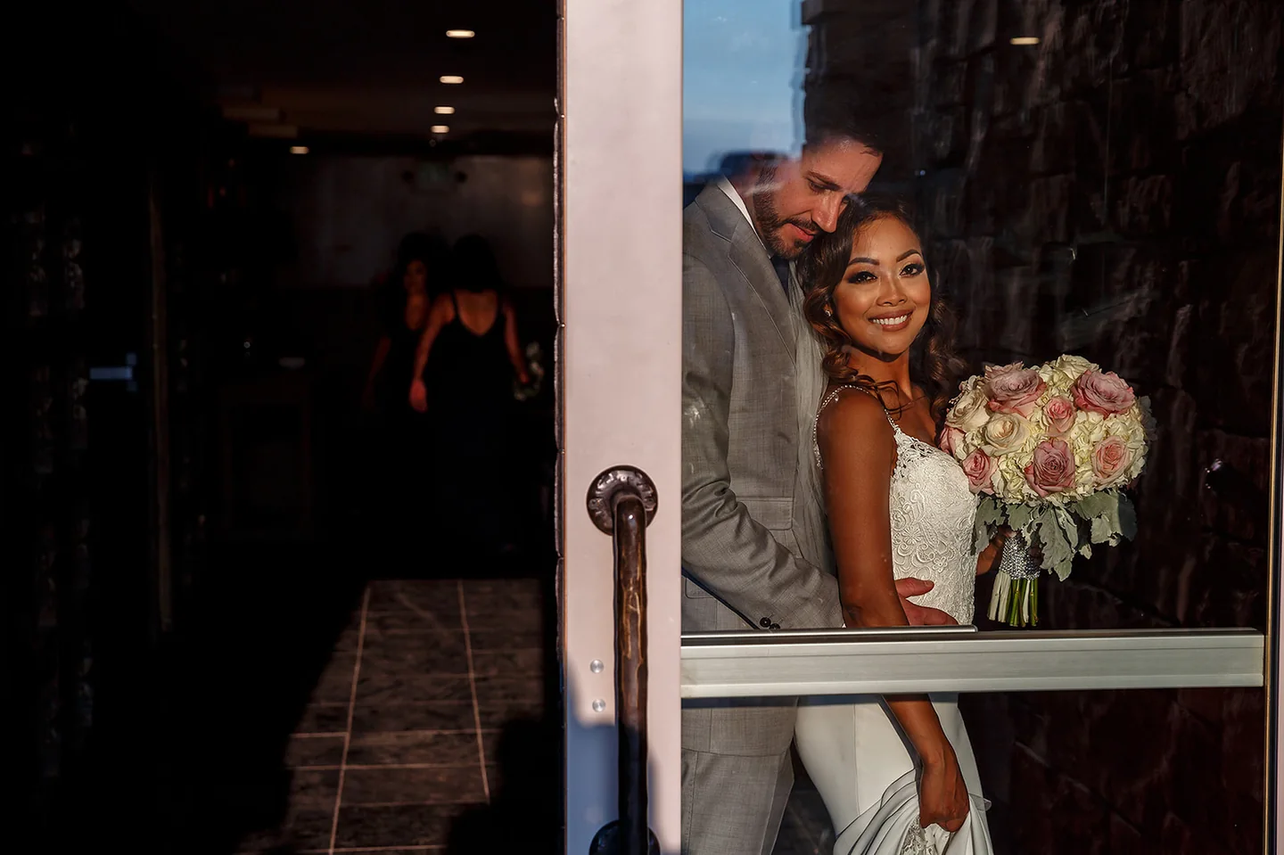 Bride holding a rose bouquet beside her groom, seen through a glass doorway