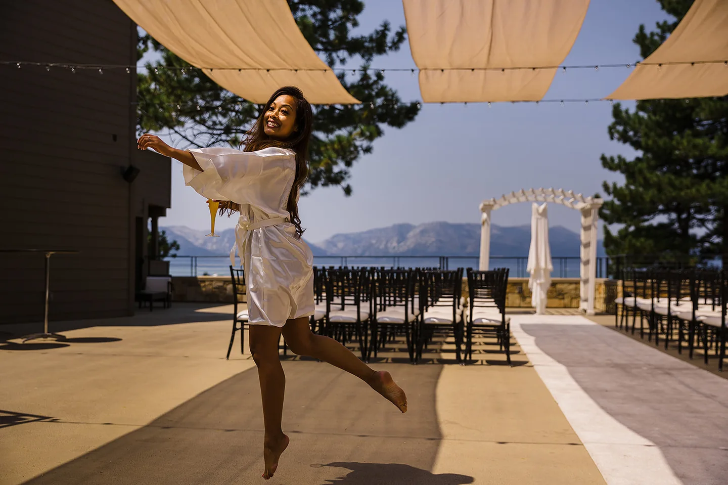 Bride jumping with joy in a white robe beside the outdoor ceremony seating