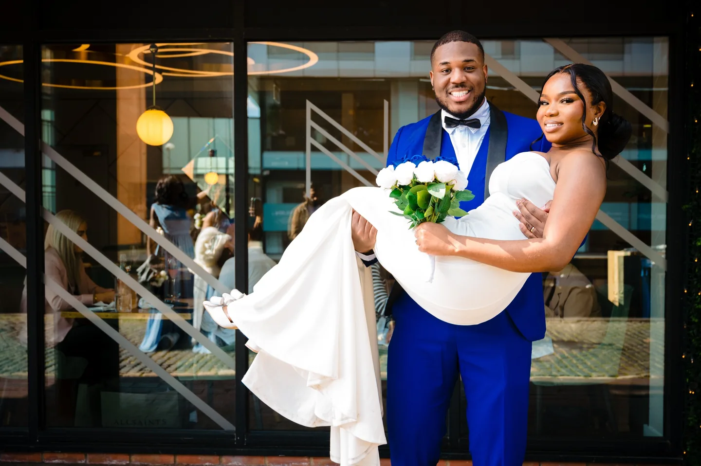 Groom in a blue suit carrying his smiling bride outside a glass-fronted building