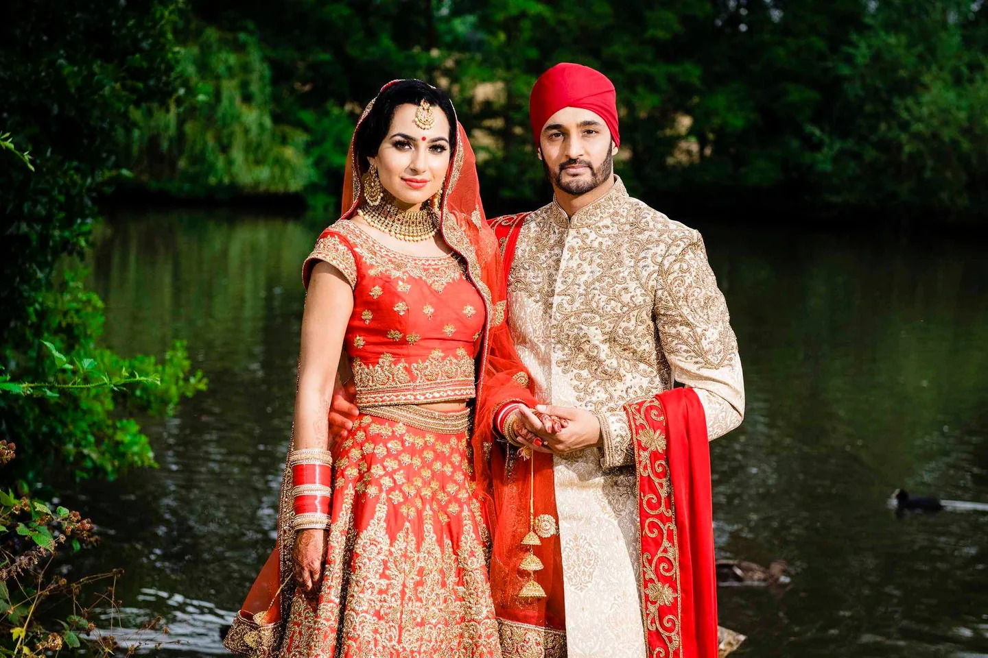 Wedding couple in red and ivory standing together beside a tree-lined pond