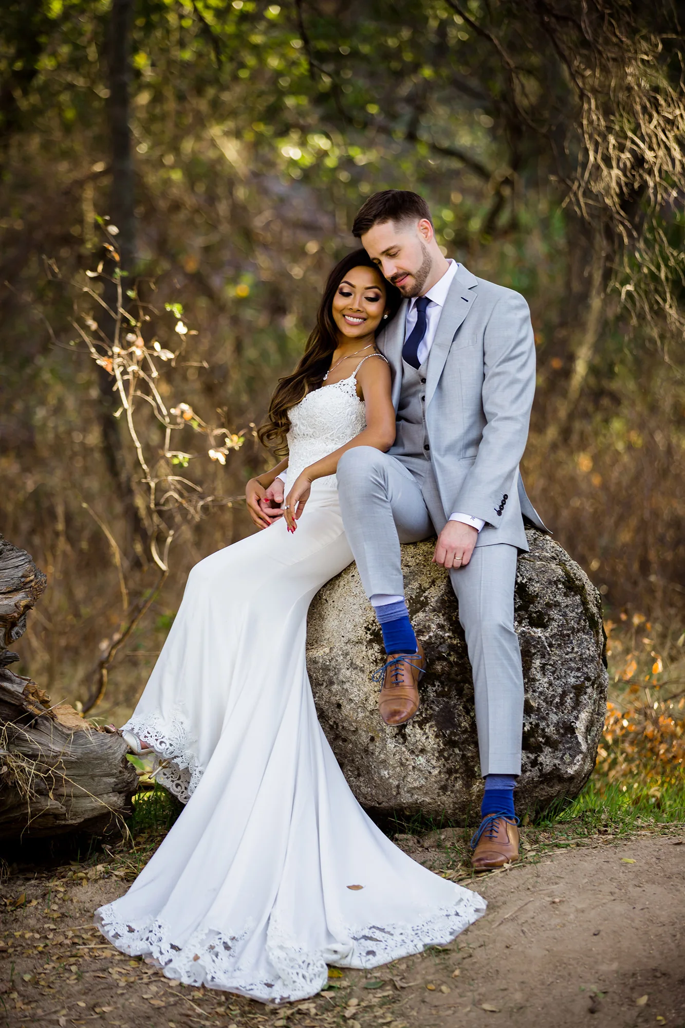 Bride and groom seated together on a woodland boulder, her dress flowing to the ground